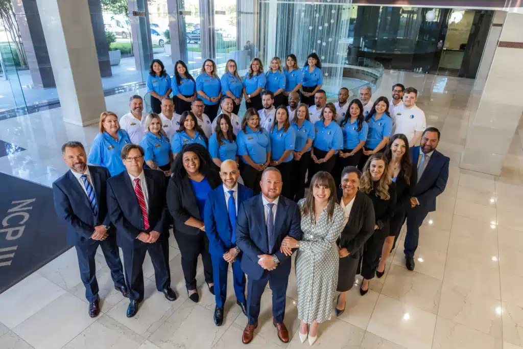 The J. Alexander Law Firm team in the lobby of the firm&rsquo;s Dallas office building, attorneys in suits in front and staff in light blue shirts behind them