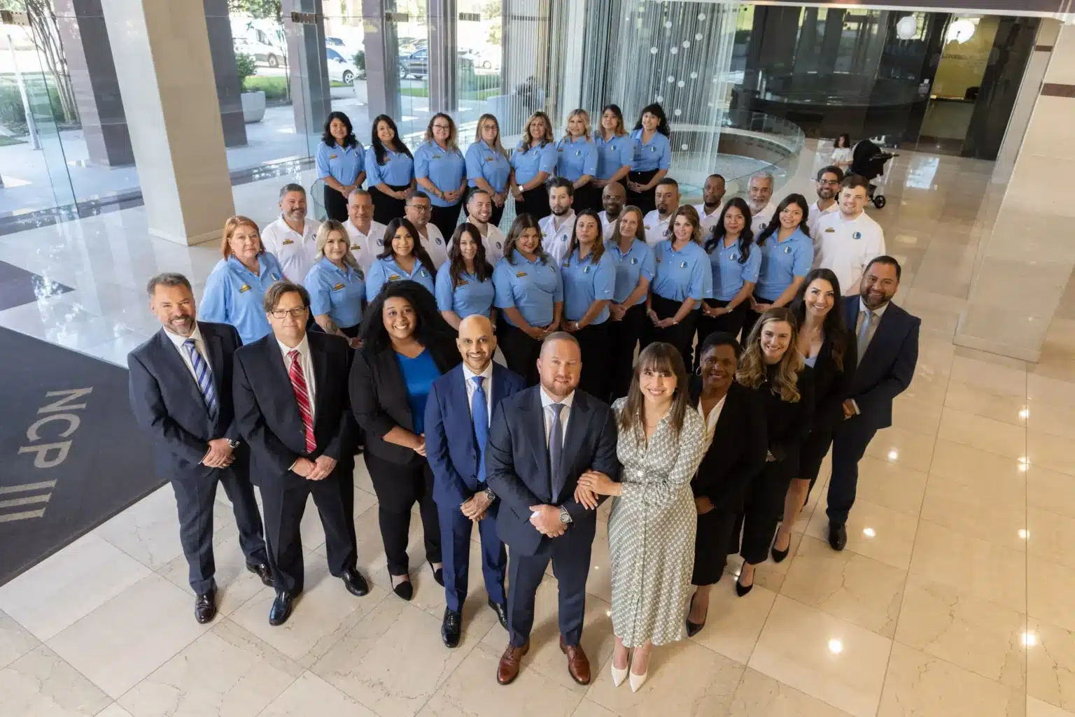 The J. Alexander Law Firm team in an office building lobby, attorneys in suits in front and staff in light blue shirts behind them