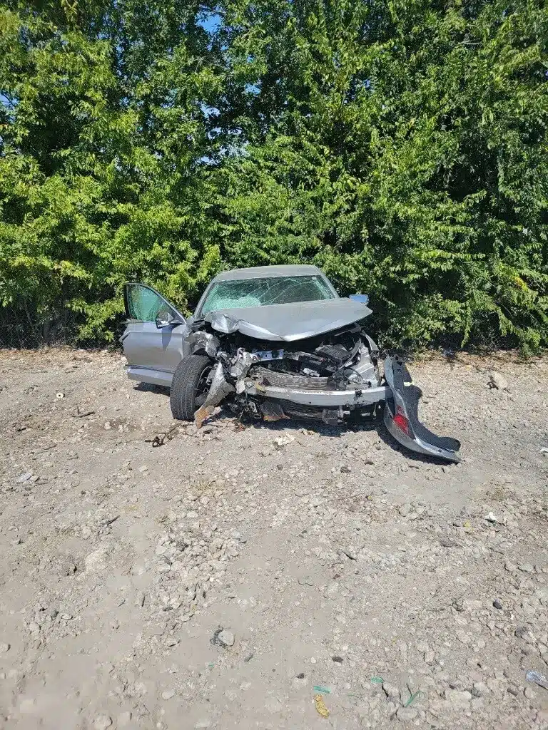 Front view of the client's silver sedan with a crushed front end, a hanging bumper cover, and a shattered windshield