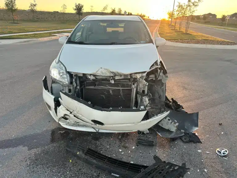 The client's white Toyota sedan stopped on Avery Nelson Parkway in Round Rock at sunrise, its front bumper torn away, hood buckled and radiator exposed, with pieces of the bumper and debris scattered across the road