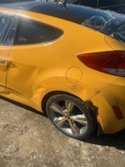 The rear passenger-side of the client's yellow hatchback parked among other cars, with the scraped quarter panel, damaged wheel area, and bumper visible