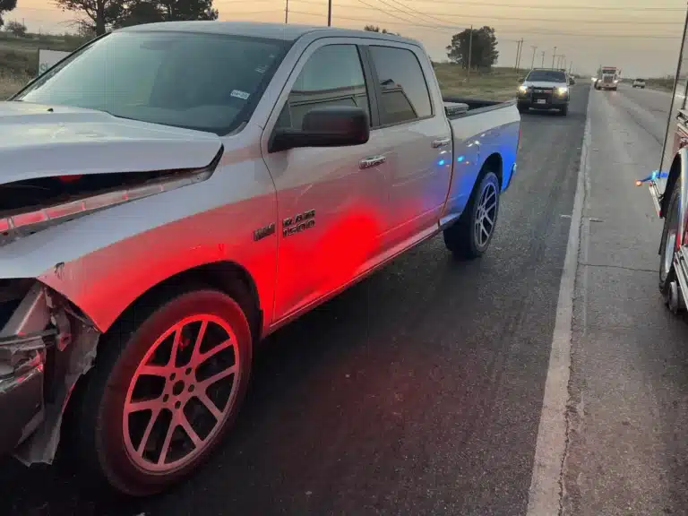 The client's silver pickup on the shoulder of FM 1788 near Midland at dawn, its hood crumpled and front bumper torn open, lit by emergency lights with traffic passing behind
