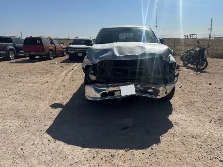 Front of the client's silver pickup in a storage lot after the FM 1788 crash near Midland, with the hood buckled, the grille pushed in and the bumper hanging loose