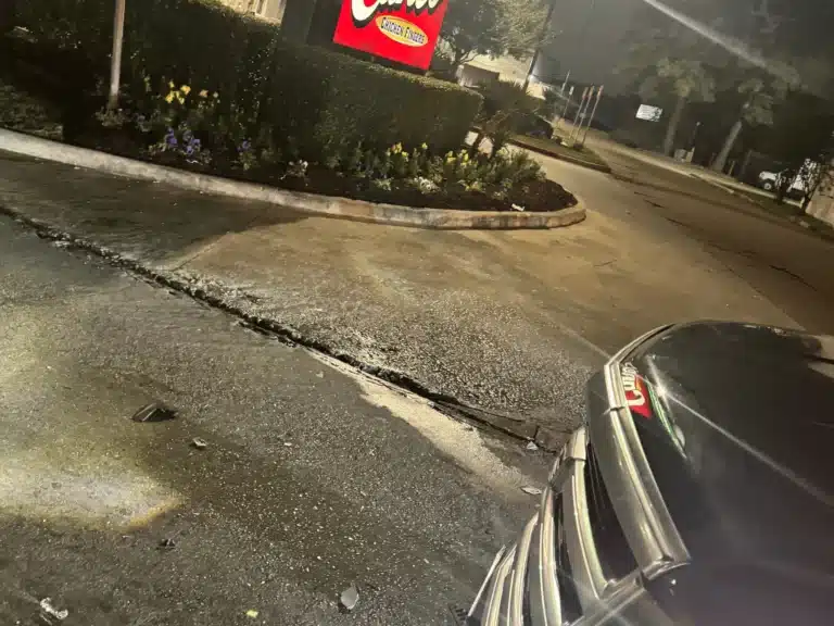 The Raising Cane’s sign and parking lot landscaping at night, with the rear of the Escalade in the foreground at the parking lot exit onto FM 1960 West