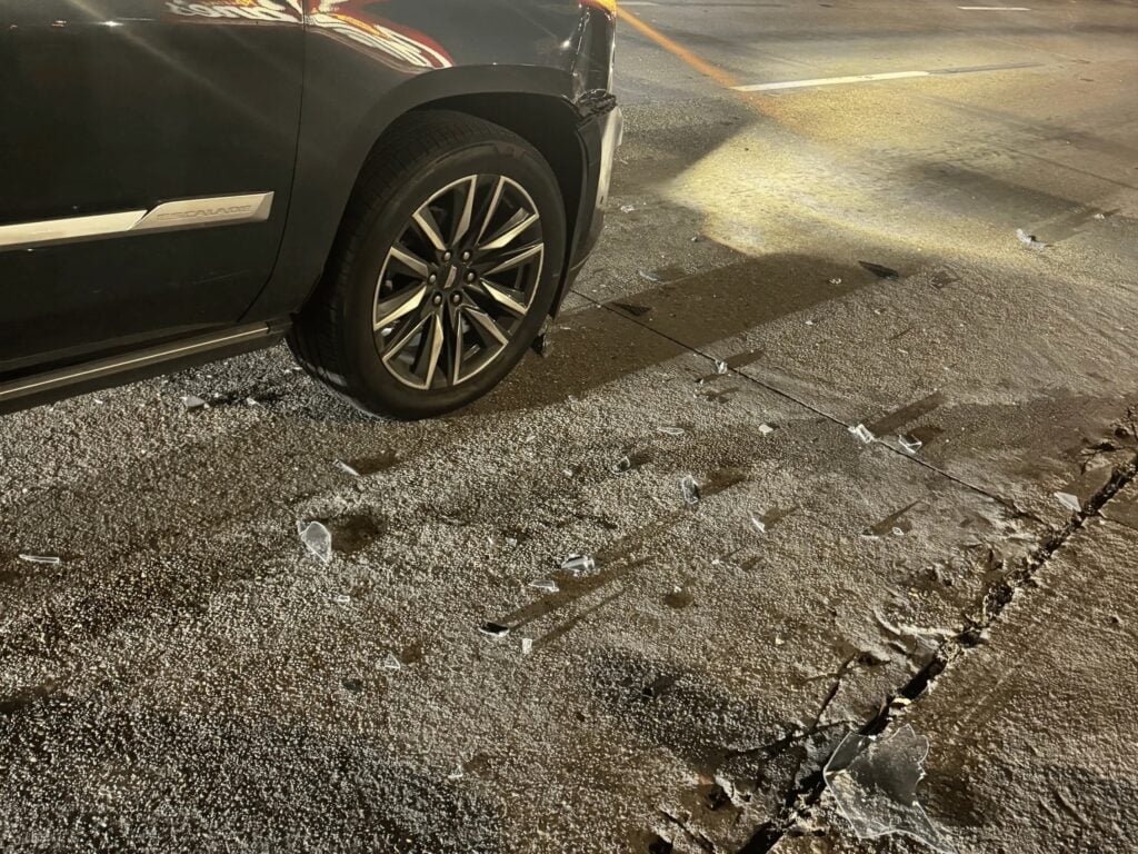 Broken vehicle pieces on the FM 1960 West pavement beside the front wheel of the client's Escalade under the streetlights