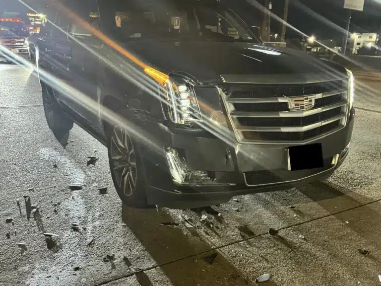 The front of the client's black Cadillac Escalade at night on FM 1960 West, with the front bumper and lower lamp housing broken and debris on the pavement