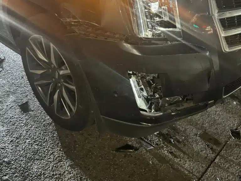 Close view of the Escalade's broken front bumper corner and lower lamp area, with broken pieces lying on the roadway below