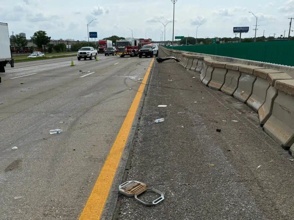Crash debris scattered across a highway lane beside the concrete barrier, with traffic stopped in the distance