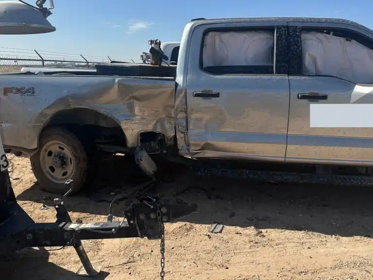 The side of the silver company pickup in a storage lot, the bed side and rear door caved in, the curtain airbags hanging, and the trailer tongue on the ground in front