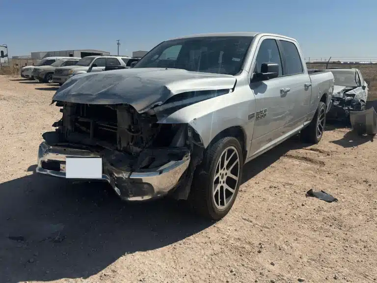 The client's silver pickup seen from the left front, with the hood folded back, the left headlight gone and the bumper cover hanging, the company truck parked behind it