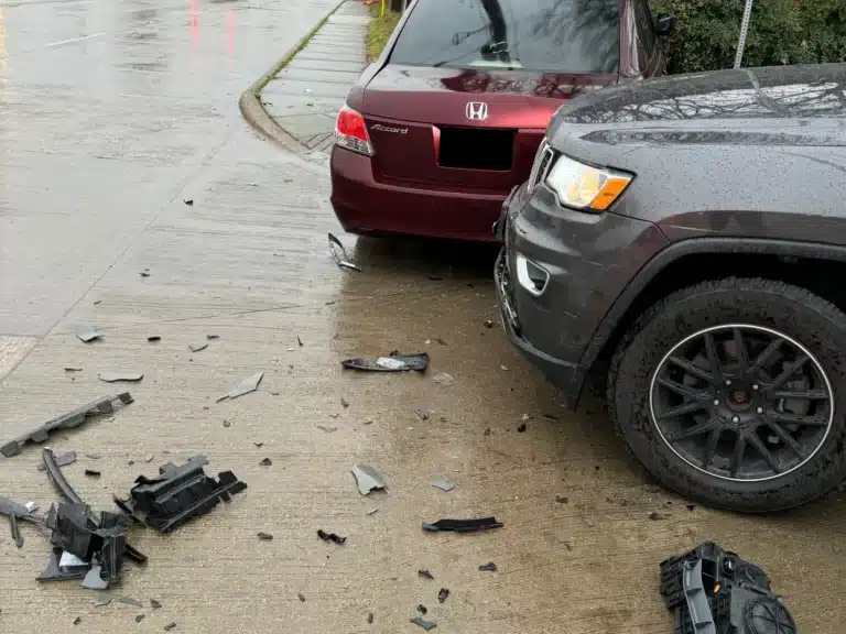 Broken car parts scattered on wet pavement between a red sedan and a dark SUV after a collision