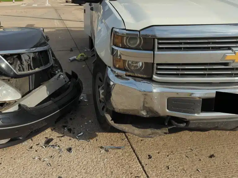 Point of impact in the Plano left turn crash: the front left area of the black Lexus against the front right bumper of the white company pickup, with broken pieces on the pavement