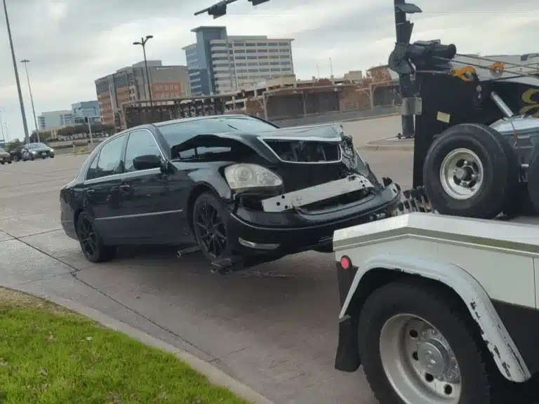 The black Lexus with a buckled hood and loose front bumper on a tow truck beside Legacy Drive after the company vehicle crash in Plano