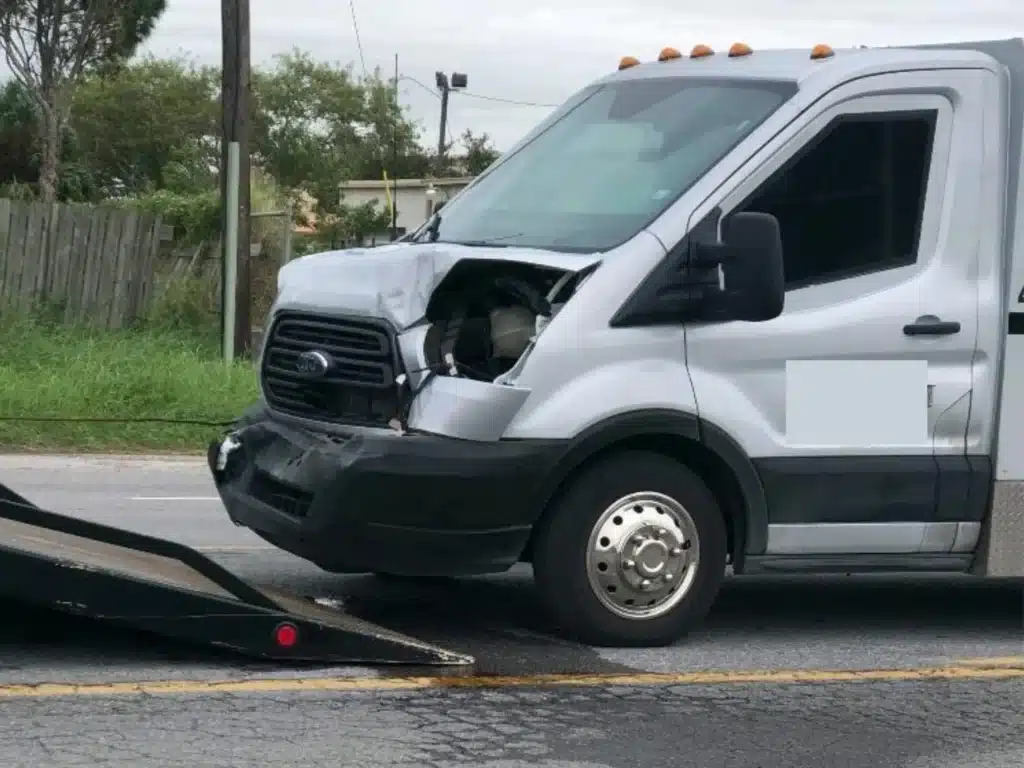 Front of the white Ford Transit armored truck with its hood buckled, the headlight housing broken open and the front bumper hanging loose, beside a tow truck bed