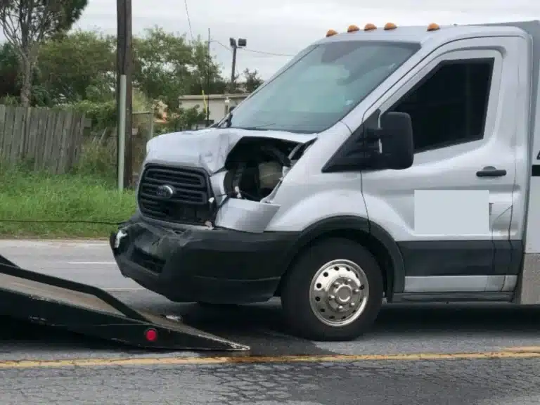 Front of the white Ford Transit armored truck from the firm's Brownsville case, its hood buckled, the headlight housing broken open and the front bumper hanging loose, beside a tow truck bed