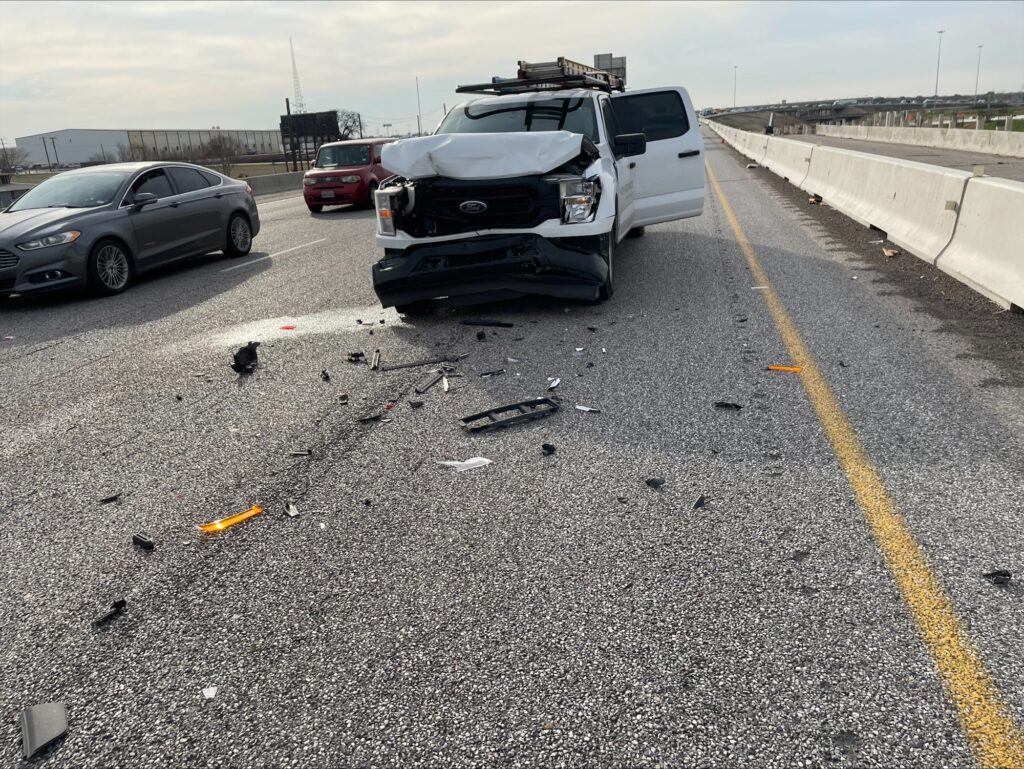 A white pickup with a crushed front end stopped on a divided highway beside the concrete barrier, with debris scattered across the lane