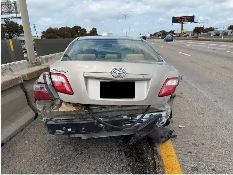 A silver Toyota Camry with a crushed rear end and its bumper hanging, stopped on a highway shoulder beside the concrete barrier