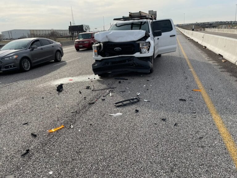 A white Ford pickup with a ladder rack and a crushed front end stopped on a highway beside the concrete barrier, with crash debris scattered across the lane