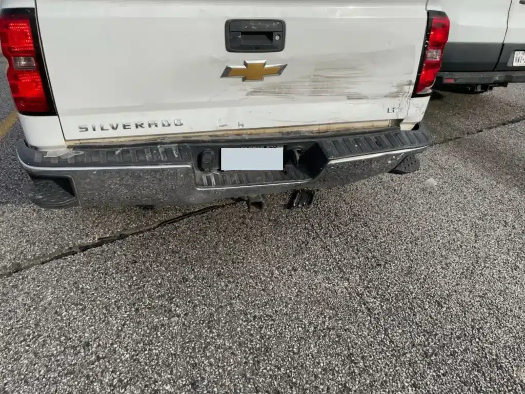 Rear view of the client's white Chevrolet Silverado showing scrapes and deformation across the tailgate and rear bumper, with the trailer hitch below