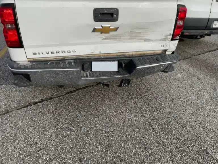 Rear view of the client's white Chevrolet Silverado showing scrapes and deformation across the tailgate and rear bumper, with the trailer hitch below