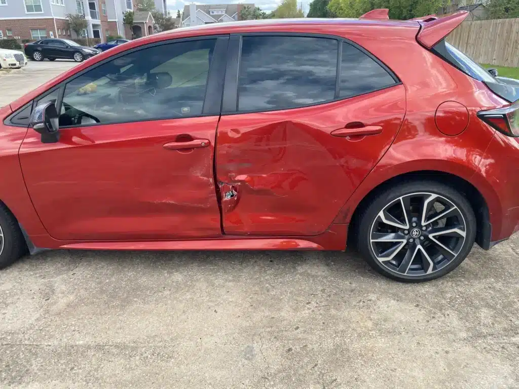 El lado del conductor del hatchback rojo de nuestra clienta, de adelante hacia atr&aacute;s, con la puerta trasera abollada