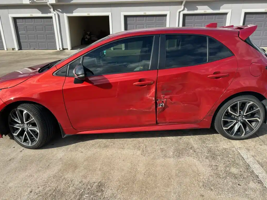 El hatchback rojo de nuestra clienta despu&eacute;s del accidente de vuelta en U en Jersey Village, con la puerta trasera del lado del conductor hundida donde la golpe&oacute; la minivan