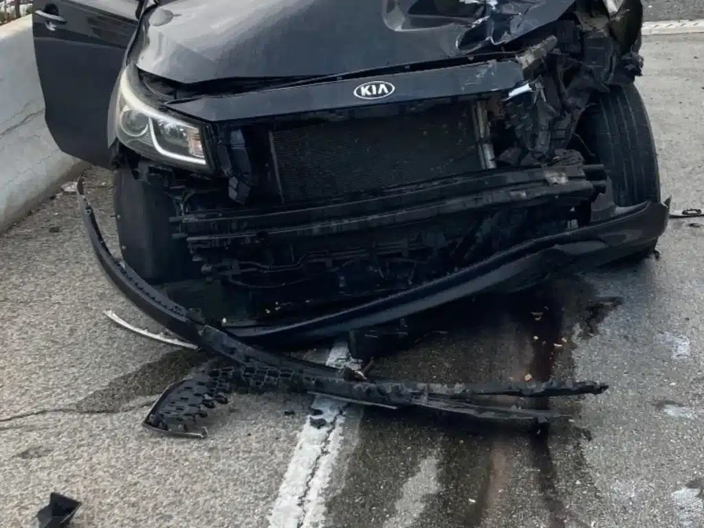 Front of the black Kia Sedona minivan that struck the RAV4, its bumper cover torn off and lying on the ramp beside a pool of leaked fluid