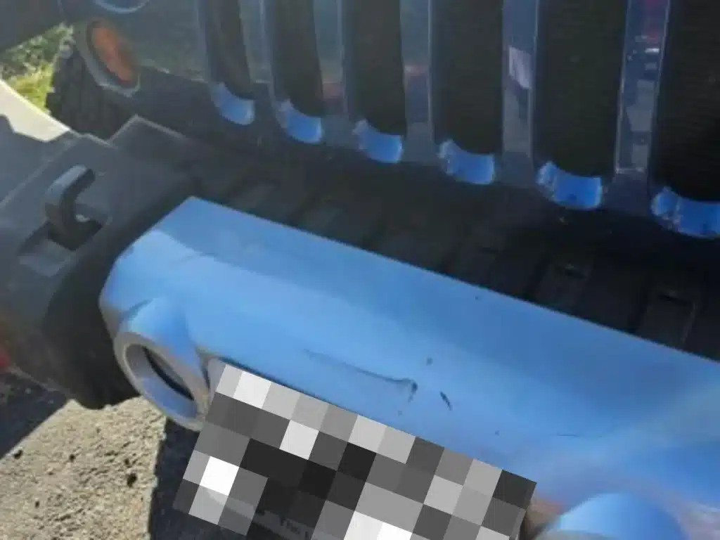 Front bumper of the blue Jeep Wrangler that rear ended the RAV4, with a crease and scuff marks beside the blurred license plate and the grille slots above