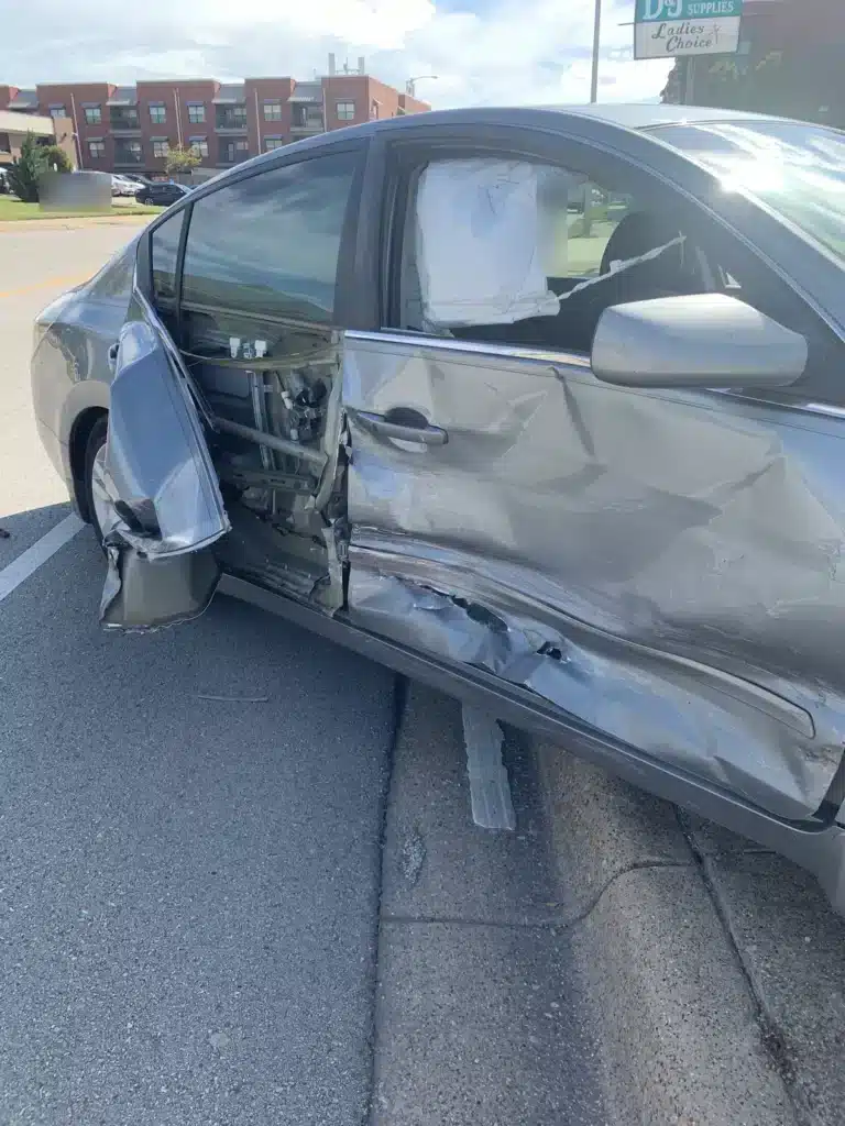 El lado del pasajero del sed&aacute;n gris de nuestra clienta despu&eacute;s del choque en la se&ntilde;al de alto, con la puerta trasera doblada y abierta, la puerta delantera hundida y el airbag lateral desplegado detr&aacute;s de la ventana