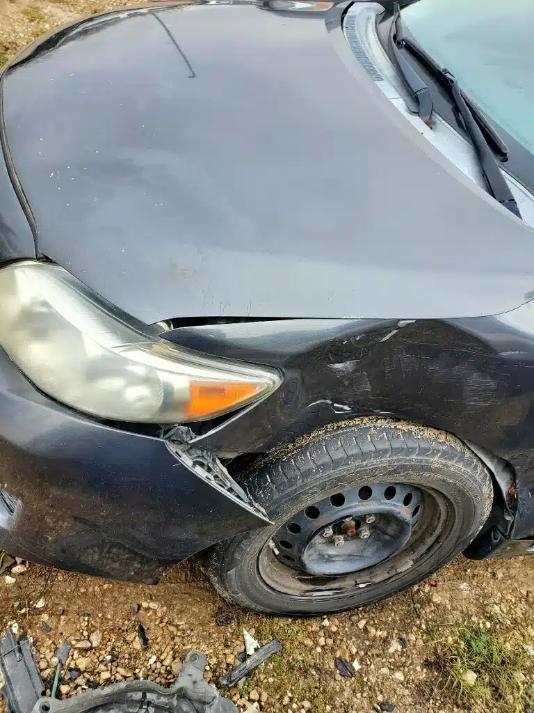 Esquina delantera de un sed&aacute;n con el faro desplazado hacia afuera, la salpicadera abollada y molduras rotas sobre grava