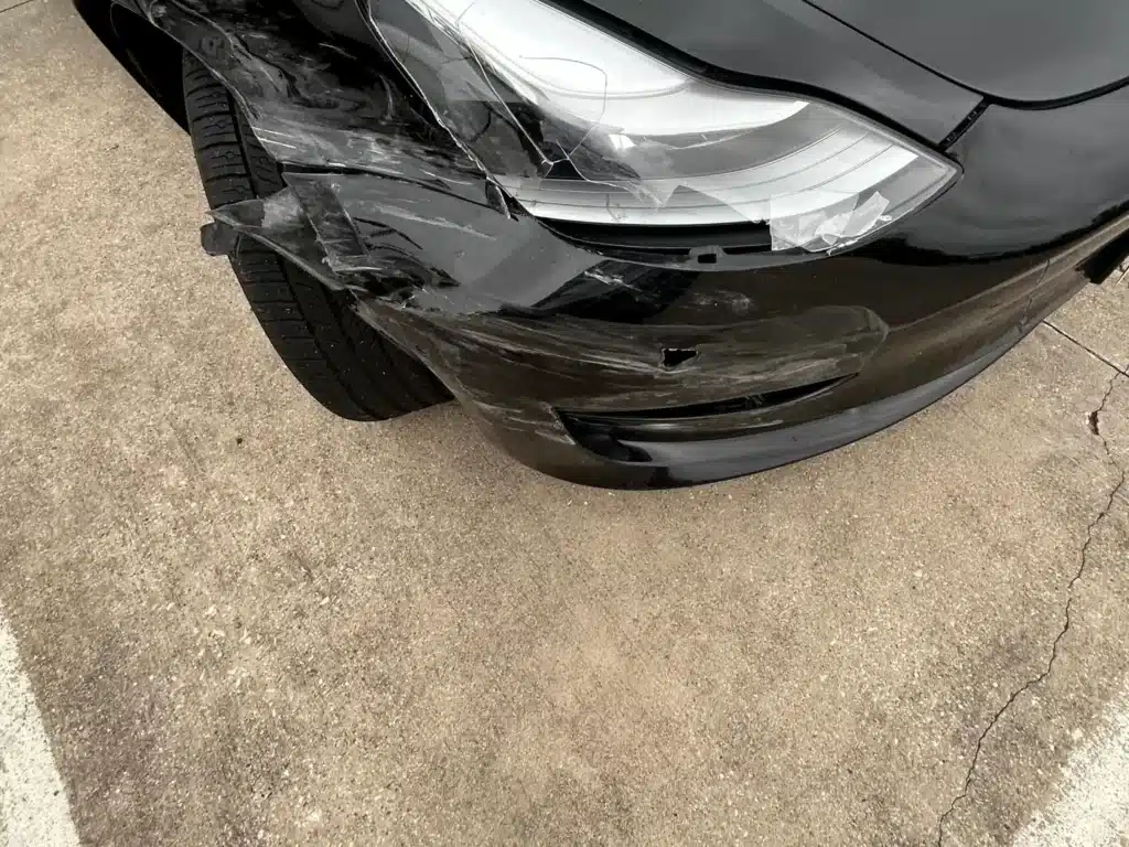Close view of the Tesla&rsquo;s front right corner, the bumper cover split and hanging and the headlamp lens shattered