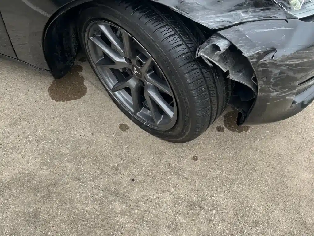 The Tesla&rsquo;s right front fender creased above the wheel, with torn bumper plastic folded into the wheel opening and fluid on the pavement