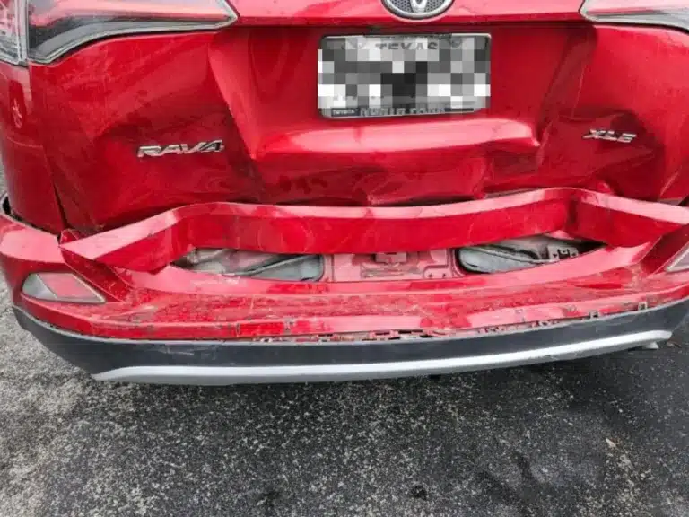 Rear of our client's red Toyota RAV4 XLE after the Jeep struck it, the bumper cover folded inward and the liftgate dented below the blurred license plate